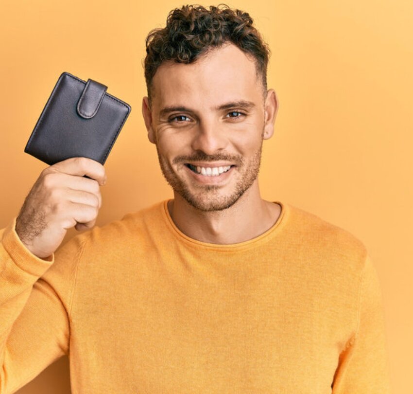 Young hispanic man holding leather wallet looking positive and happy standing and smiling with a confident smile showing teeth