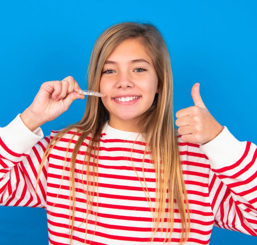 caucasian teen girl wearing striped shirt over blue studio background holding an invisible braces aligner and rising thumb up, recommending this new treatment. Dental healthcare concept.
